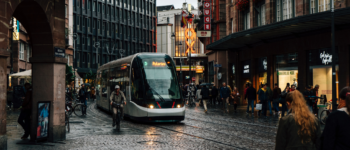 Photographie d'un tram arrivant vers la place de l'homme de fer à strasbourg. on voit le cinéma vox en arrière plan, et des cyclistes et piétons sur le côté.