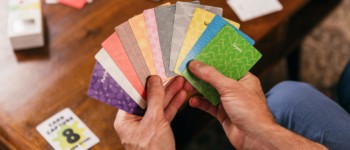 mains tenant des cartes de différentes couleurs au dessus d'une table où sont posées d'autres cartes